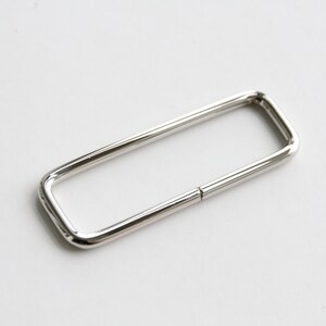 May include: A silver metal rectangular ring with a smooth finish. The ring is open on one side and has a slight bend in the middle.