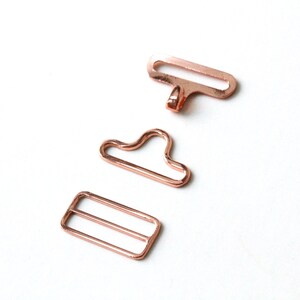 May include: Three rose gold metal clasps for sewing projects. The clasps are in different shapes, including a rectangle, a loop, and a hook.