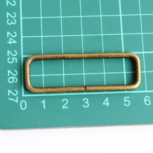 May include: A gold-colored metal rectangular loop with rounded edges. The loop is on a green cutting mat with white numbers.