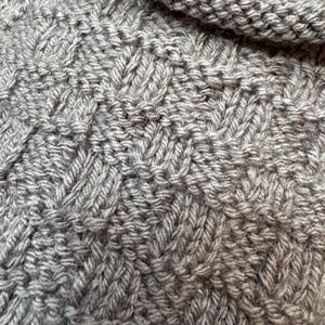 Pu&ograve; includere: Una vista ravvicinata di un tessuto a maglia color tortora caratterizzato da un motivo ripetitivo a intreccio. Il tessuto presenta una superficie strutturata creata dall'alternanza di punti a maglia verticali e orizzontali, risultando in un aspetto denso e uniforme.