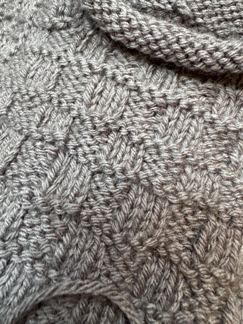 Pu&ograve; includere: Una vista ravvicinata di un tessuto a maglia color tortora caratterizzato da un motivo ripetitivo a intreccio. Il tessuto presenta una superficie strutturata creata dall'alternanza di punti a maglia verticali e orizzontali, risultando in un aspetto denso e uniforme.