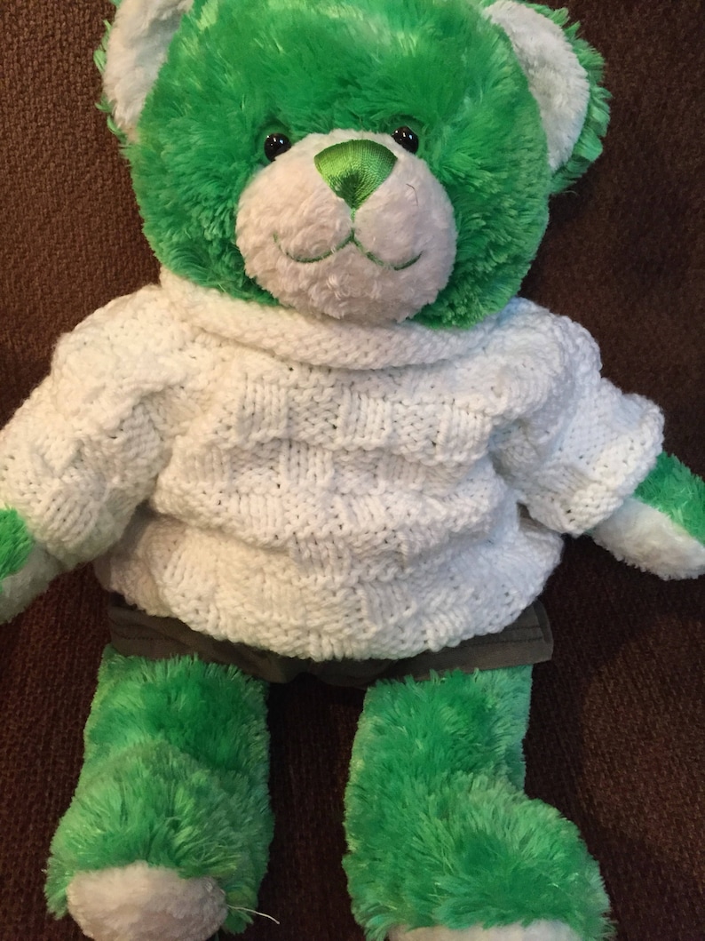 Pu&ograve; includere: Un orsacchiotto di peluche con pelliccia verde brillante e bianca, che indossa un maglione di maglia bianco con un motivo a intreccio testurizzato e pantaloncini grigio scuro. L'orso ha un naso in raso verde, occhi in plastica nera e zampe bianche.
