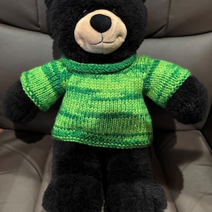 May include: A black plush bear wearing a green and light green striped sweater. The bear has a tan muzzle and a smiling expression. The bear is sitting on a gray surface.