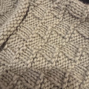 Pu&ograve; includere: Un tessuto a maglia beige chiaro caratterizzato da un motivo ripetitivo a intreccio. Il tessuto &egrave; realizzato con filato spesso, creando una superficie strutturata con quadrati alternati di punti verticali e orizzontali a griglia.