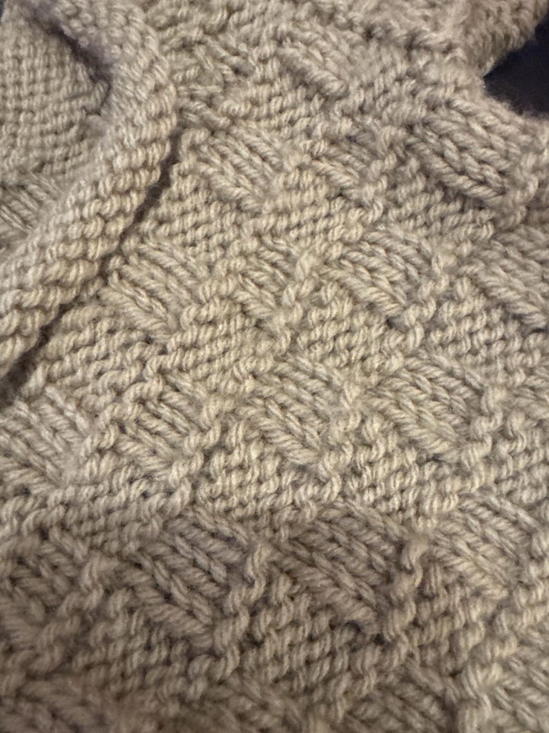 Pu&ograve; includere: Un tessuto a maglia beige chiaro caratterizzato da un motivo ripetitivo a intreccio. Il tessuto &egrave; realizzato con filato spesso, creando una superficie strutturata con quadrati alternati di punti verticali e orizzontali a griglia.