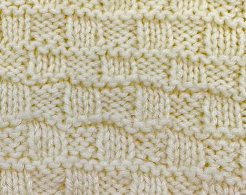 Pu&ograve; includere: Una vista ravvicinata di un tessuto lavorato a maglia color crema con un motivo a intreccio testurizzato. Il design &egrave; costituito da quadrati alternati di punti a maglia rasata verticali e punti a legaccio orizzontali, creando un effetto a griglia.