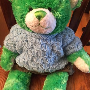 Pu&ograve; includere: Un orsacchiotto di peluche con pelliccia verde brillante, muso bianco e naso cucito verde. L'orso indossa un maglione lavorato a maglia azzurro con un motivo a intreccio testurizzato e pantaloncini grigio scuro. L'orso ha cuscinetti bianchi.