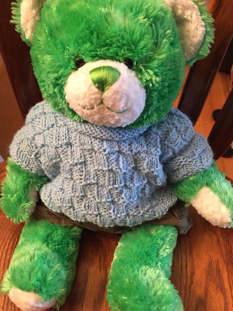 Pu&ograve; includere: Un orsacchiotto di peluche con pelliccia verde brillante, muso bianco e naso cucito verde. L'orso indossa un maglione lavorato a maglia azzurro con un motivo a intreccio testurizzato e pantaloncini grigio scuro. L'orso ha cuscinetti bianchi.