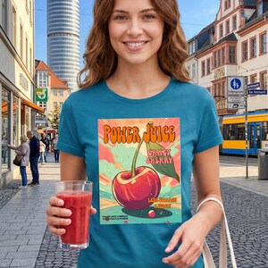 May include: A teal short-sleeve t-shirt featuring a graphic print of a red cherry with a green leaf. The text reads Power Juice, Crazy Cherry, and includes smaller text at the bottom. The design has a colorful background with sunburst rays.