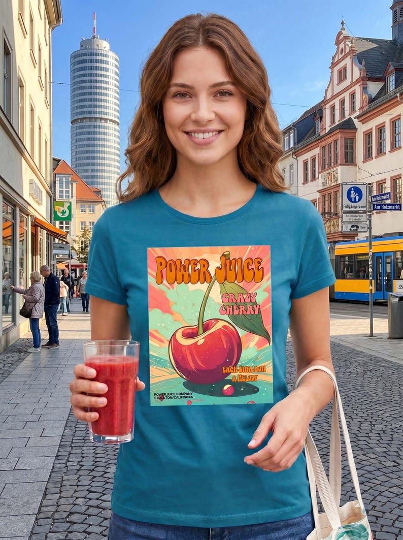 May include: A teal short-sleeve t-shirt featuring a graphic print of a red cherry with a green leaf. The text reads Power Juice, Crazy Cherry, and includes smaller text at the bottom. The design has a colorful background with sunburst rays.