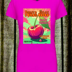 May include: A bright fuchsia short-sleeved t-shirt featuring a central graphic of a red cherry with a green leaf. The text reads POWER JUICE, CRAZY CHERRY, LACK SHALLOW A MELODY, and POWER JUICE COMPANY STOCKTON CALIFORNIA against a colorful background.