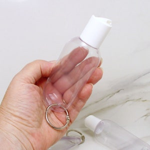 May include: A clear plastic bottle with a white cap and a silver key ring. The bottle is designed to hold hand sanitizer or other liquids.