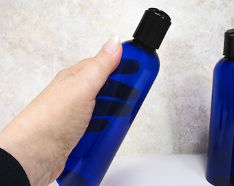 May include: A cobalt blue bottle with a black cap is held in a hand. The bottle has a sleek, cylindrical shape and appears to be made of glass or a similar material. The background is a neutral, off-white color.
