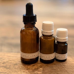 May include: Three amber glass bottles of essential oils with white labels. The largest bottle has a black dropper, and the labels read "COCONUT VANILLA", "ORCHID+LILY", and "MAPLE L". The bottles are arranged on a wooden surface.