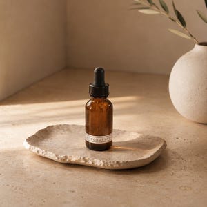 May include: A small amber glass dropper bottle with a black cap and a white label reading COCONUT VANILLA, resting on a textured, light-colored, irregular stone tray. The bottle contains a liquid and features a glass pipette dropper mechanism.