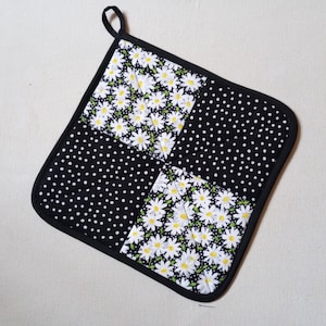 May include: Black and white quilted hot pad with a daisy print and polka dot pattern. The hot pad has a black fabric border.