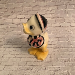 May include: A ceramic duck figurine wearing a pink and black striped scarf and a black and white hat. The duck is standing on a white brick background.