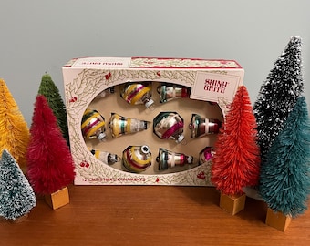 May include: A box of 12 Shiny Brite Christmas ornaments. The ornaments are in a variety of shapes and colors, including red, green, blue, and gold. The box is decorated with a holly and berry design. The text "Shiny Brite" is printed on the box.
