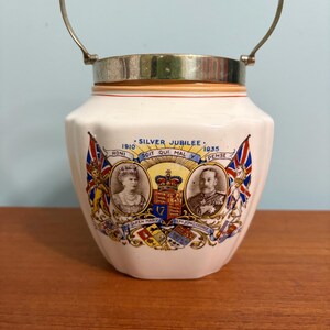 May include: White ceramic jar with a silver lid and handle. The jar features a colourful image of King George V and Queen Mary, commemorating their Silver Jubilee in 1935. The text "1910 SILVER JUBILEE 1935" is printed on the jar.
