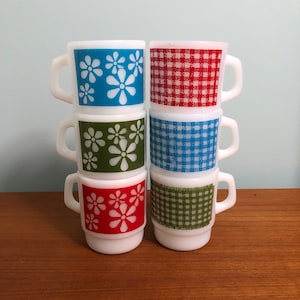 May include: Six white ceramic mugs stacked in a pyramid. The mugs have different patterns: blue with white flowers, red gingham, green with white flowers, blue gingham, green gingham, and red with white flowers.