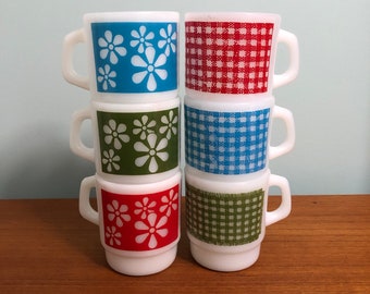 May include: Six white ceramic mugs stacked in a pyramid. The mugs have different patterns: blue with white flowers, red gingham, green with white flowers, blue gingham, green gingham, and red with white flowers.