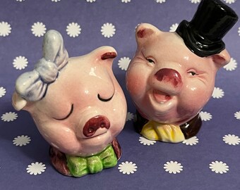 Könnte beinhalten: Ein Paar Salz- und Pfefferstreuer aus Keramik in Form von Schweinen. Das weibliche Schwein trägt eine blaue Schleife und eine grüne Fliege. Das männliche Schwein trägt einen schwarzen Zylinder und eine gelbe Fliege.