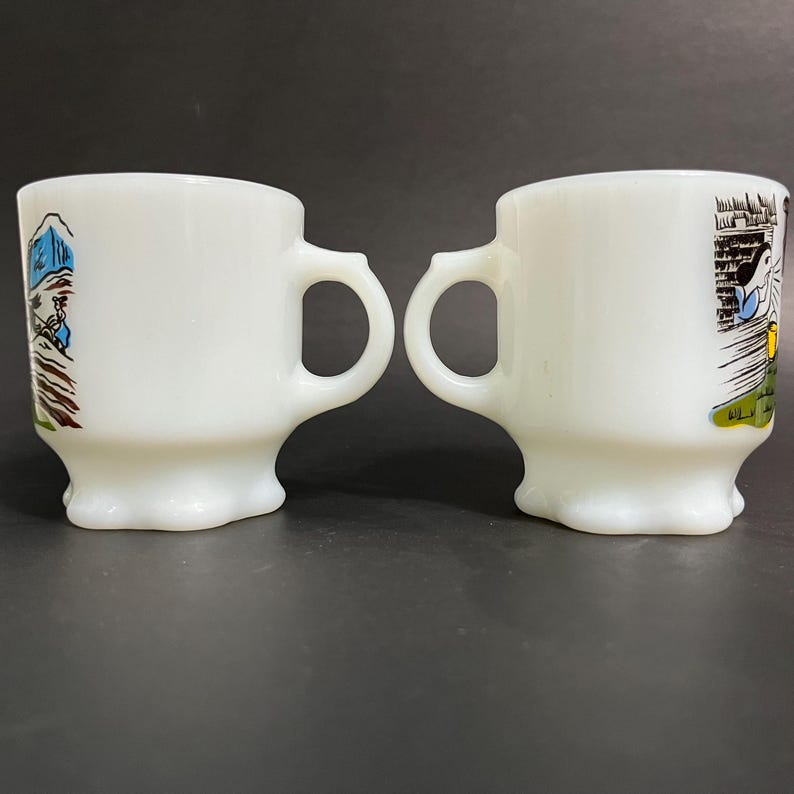 May include: Two white ceramic mugs with a footed base and a handle. One mug has a colorful illustration of a mountain scene, the other has a colorful illustration of a building with a woman in a blue dress standing in front of it.