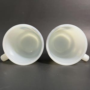 May include: Two white ceramic mugs with a smooth, glossy finish. The mugs have a rounded shape and a handle on one side.