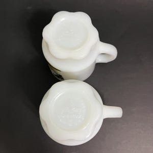 May include: Two white milk glass mugs with a scalloped rim and a floral design. The mugs are made by Fire King and have the words "Fire King" and "Made in USA" on the bottom.