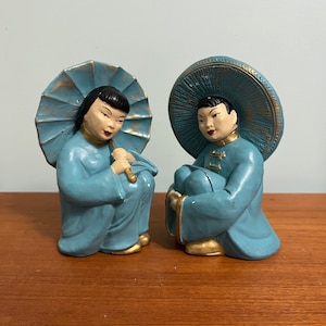 Peut inclure: Deux figurines en céramique bleue représentant des personnes vêtues de vêtements traditionnels asiatiques. La figurine de gauche tient un parapluie bleu et or. La figurine de droite porte un chapeau bleu et or.