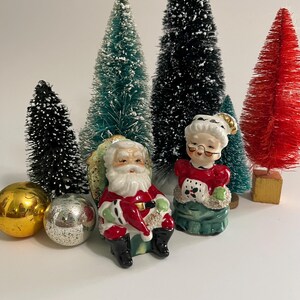 K&ouml;nnte beinhalten: Vintage Salz- und Pfefferstreuer aus Keramik mit Weihnachtsmann und Frau Claus. Der Weihnachtsmann tr&auml;gt einen roten Anzug mit wei&szlig;en Verzierungen und einen wei&szlig;en Bart. Frau Claus tr&auml;gt ein rotes Kleid mit wei&szlig;en Verzierungen und eine wei&szlig;e Sch&uuml;rze. Beide sitzen auf gr&uuml;nen Sockeln.