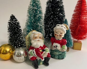 May include: Vintage ceramic Santa Claus and Mrs. Claus salt and pepper shakers. Santa is wearing a red suit with white trim and a white beard. Mrs. Claus is wearing a red dress with white trim and a white apron. They are both sitting on green bases.