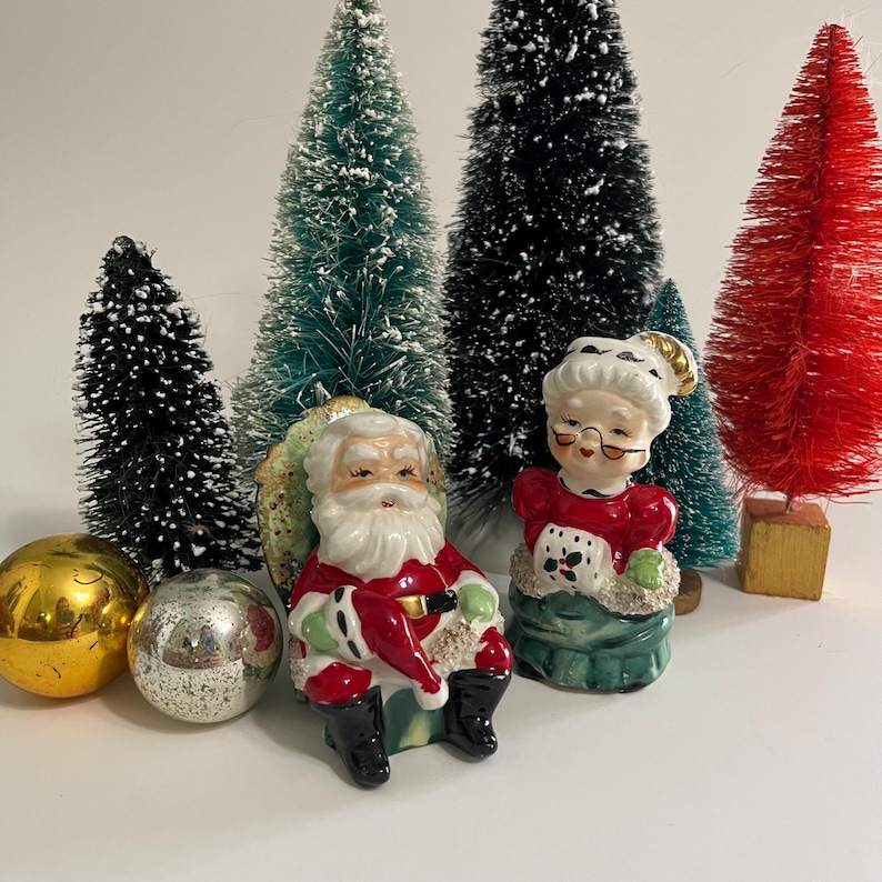K&ouml;nnte beinhalten: Vintage Salz- und Pfefferstreuer aus Keramik mit Weihnachtsmann und Frau Claus. Der Weihnachtsmann tr&auml;gt einen roten Anzug mit wei&szlig;en Verzierungen und einen wei&szlig;en Bart. Frau Claus tr&auml;gt ein rotes Kleid mit wei&szlig;en Verzierungen und eine wei&szlig;e Sch&uuml;rze. Beide sitzen auf gr&uuml;nen Sockeln.