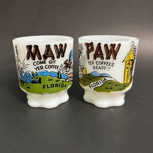 Peut inclure: Deux tasses en céramique blanche sur le thème de la Floride vintage. La tasse de gauche dit "Maw Come Git Yer Coffee!" et la tasse de droite dit "Paw Yer Coffee's Ready!!". Les deux tasses ont une illustration verte, bleue et brune d'un paysage de Floride.