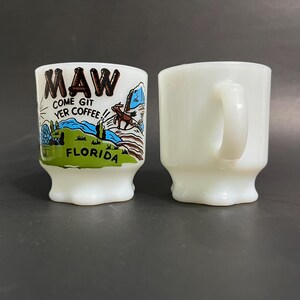 May include: Two white ceramic mugs with a blue and green design. The mug on the left has the text "Maw Come Git Yer Coffee! Florida".