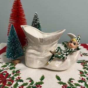 May include: A white ceramic Christmas boot planter with a green and white elf figurine sitting on the edge. The boot has a pearlescent finish.
