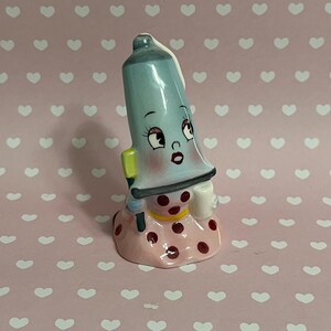 Peut inclure: Une figurine en céramique représentant un tube de dentifrice de dessin animé portant une robe rose à pois et tenant une brosse à dents. Le tube a un corps bleu et blanc avec un visage souriant et de grands yeux.