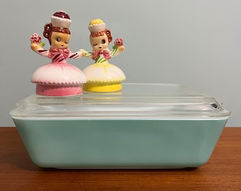May include: A vintage turquoise blue glass baking dish with a clear glass lid. Two ceramic figurines of girls in dresses are on top of the lid. The girls are wearing pink and yellow dresses with flowers.