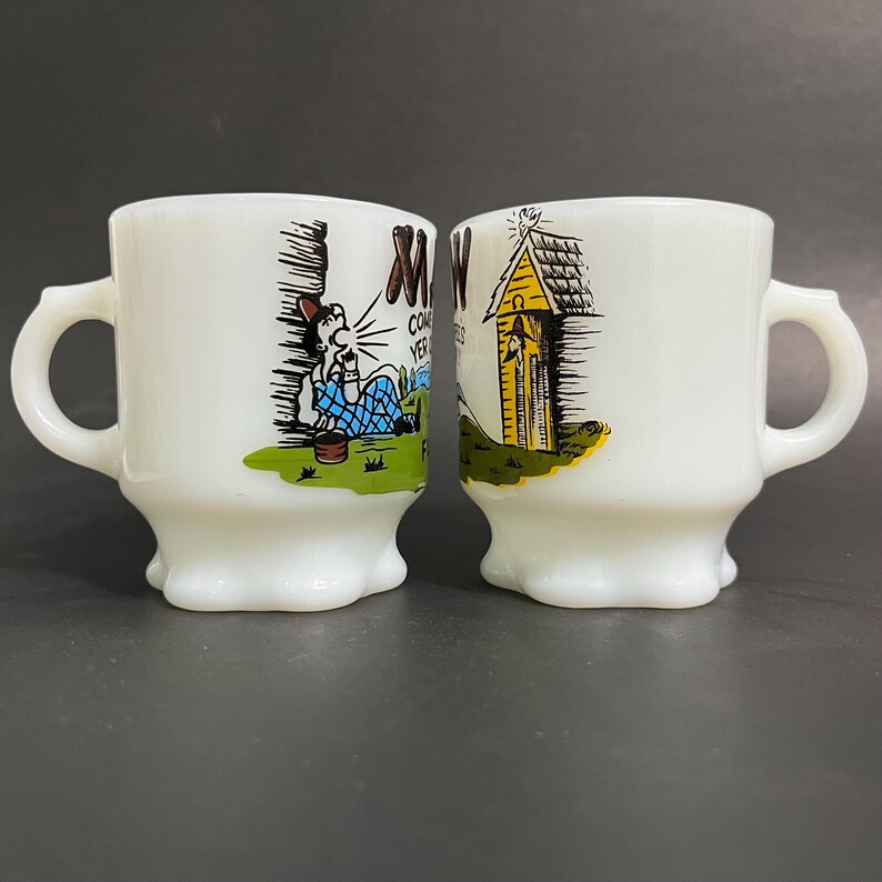 May include: Two white milk glass mugs with cartoon illustrations. One mug features a man sitting on a hill with the text "Come Yer" and the other mug features a yellow outhouse with a bell on top.