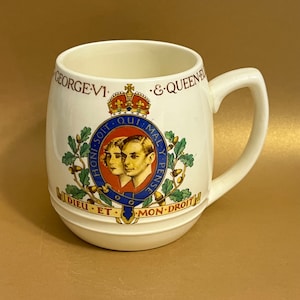 May include: White ceramic mug with a royal crest featuring King George VI and Queen Elizabeth. The crest is surrounded by oak leaves and acorns. The mug has the text "GEORGE VI & QUEEN ELIZA" and "DIEU ET MON DROIT" in gold lettering.