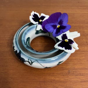 May include: A white ceramic ring vase with blue and green glaze and black accents. The vase is stacked with two rings and has three purple and white pansies in it.