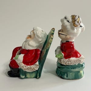 K&ouml;nnte beinhalten: Ein vintage Keramik-Salz- und Pfefferstreuer-Set mit Weihnachtsmann und Frau Claus. Der Weihnachtsmann sitzt in einem gr&uuml;nen Schaukelstuhl mit wei&szlig;em Bart und rotem Anzug. Frau Claus tr&auml;gt ein rotes Kleid mit wei&szlig;em Kragen und gr&uuml;nem Rock. Beide Figuren haben einen wei&szlig;en und goldenen Besatz.
