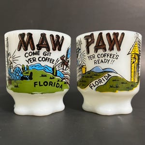 May include: Two white ceramic mugs with a vintage design. The mugs are decorated with a cartoon scene of a Florida landscape. One mug says "Maw Come Git Yer Coffee!" and the other says "Paw Yer Coffee's Ready!!".