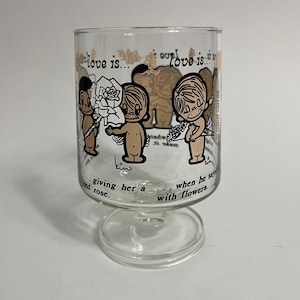 May include: A clear glass goblet with a stem and a base. The goblet has a cartoon illustration of two couples with the text "love is... love is... giving her a red rose... when he says with flowers."
