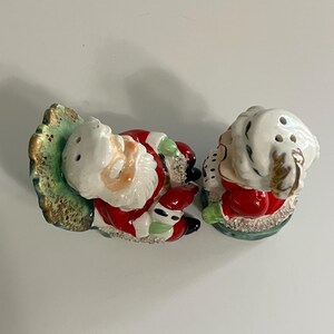 K&ouml;nnte beinhalten: Vintage Salz- und Pfefferstreuer aus Keramik mit Santa Claus und Mrs. Claus, die in Schaukelst&uuml;hlen sitzen. Die Streuer sind in Rot, Wei&szlig; und Gr&uuml;n mit goldenen Akzenten bemalt.