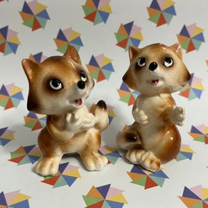 May include: Two ceramic raccoon figurines, one with its paws clasped in front of its chest and the other with one paw raised. Both raccoons have brown fur, black eyes, and white noses.