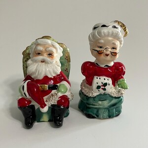 K&ouml;nnte beinhalten: Ein vintage Keramik-Salz- und Pfefferstreuer-Set mit Weihnachtsmann und Frau Claus. Der Weihnachtsmann sitzt auf einem gr&uuml;nen Stuhl mit einem rot-wei&szlig;en Anzug, wei&szlig;em Bart und schwarzen Stiefeln. Frau Claus tr&auml;gt ein rotes Kleid mit einer wei&szlig;en Sch&uuml;rze und einem gr&uuml;nen Rock. Sie tr&auml;gt einen wei&szlig;en Hut mit schwarzem Besatz und eine schwarze Brille.