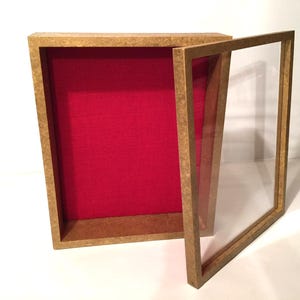 May include: A rectangular shadow box display case with a textured light brown frame and a vibrant red fabric-lined interior. The case features a removable front frame section with a clear glass panel, allowing for the display of various items.