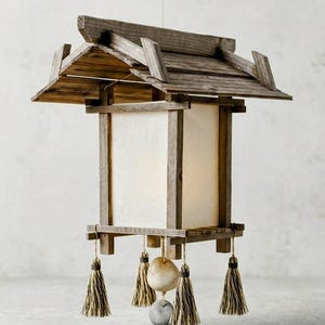 May include: A hanging lantern featuring a weathered wooden frame with a gabled roof structure and a central white translucent panel. Four decorative tassels hang from the corners, while a central cord suspends two smooth, rounded stones.