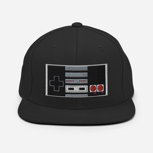 May include: Black and grey baseball cap with an embroidered image of a classic video game controller. The controller has red buttons and the text "Select Start" is visible.
